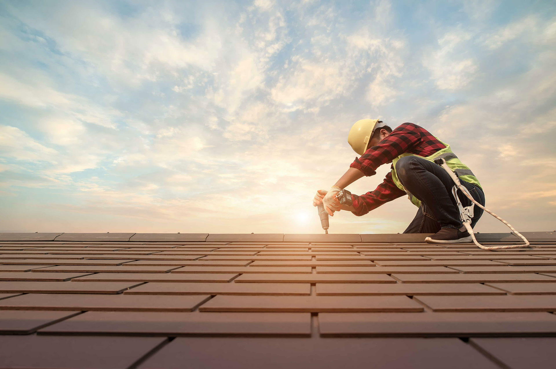 The image shows a construction worker kneeling on a rooftop during sunset, with a focus on the worker s hands holding a tool.